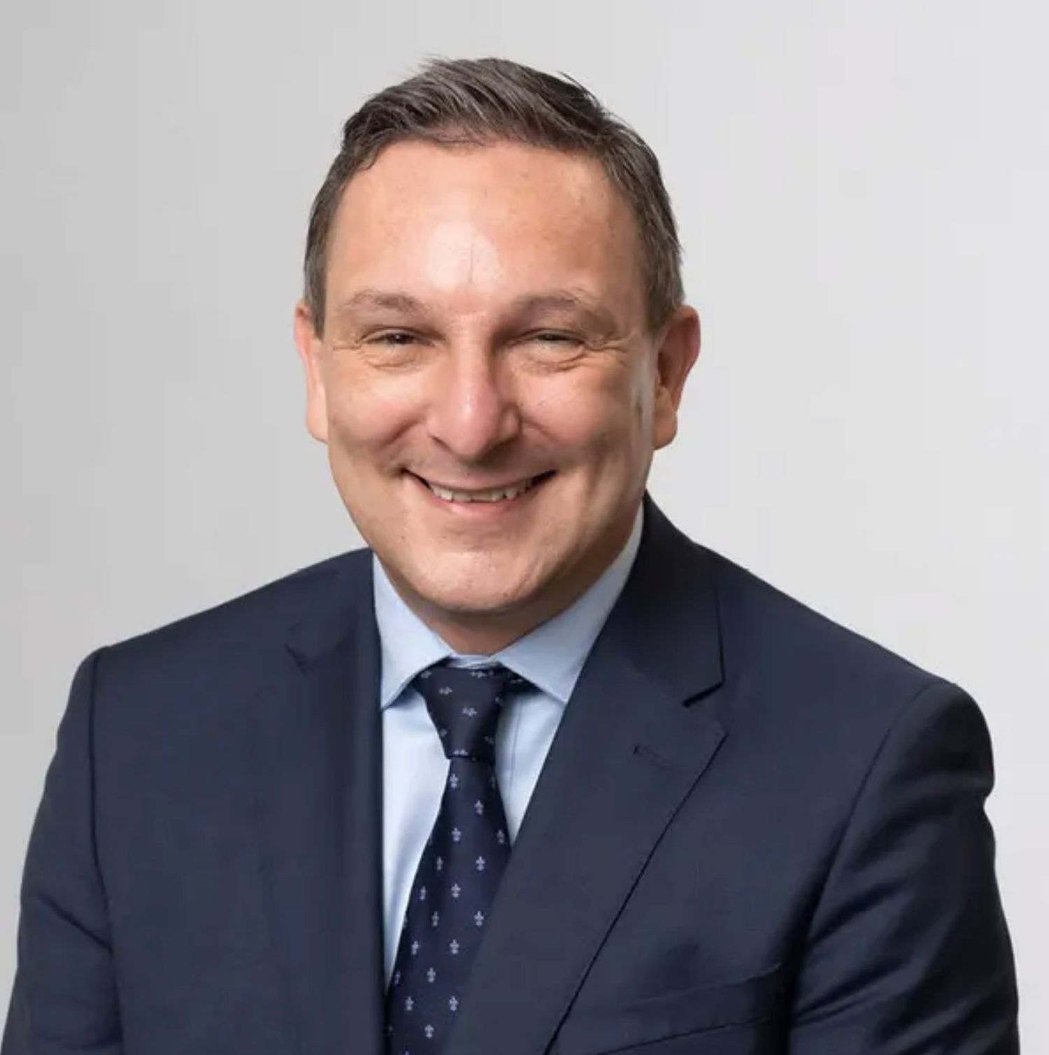 Professional businessman in navy suit and tie smiling confidently against a light gray background.