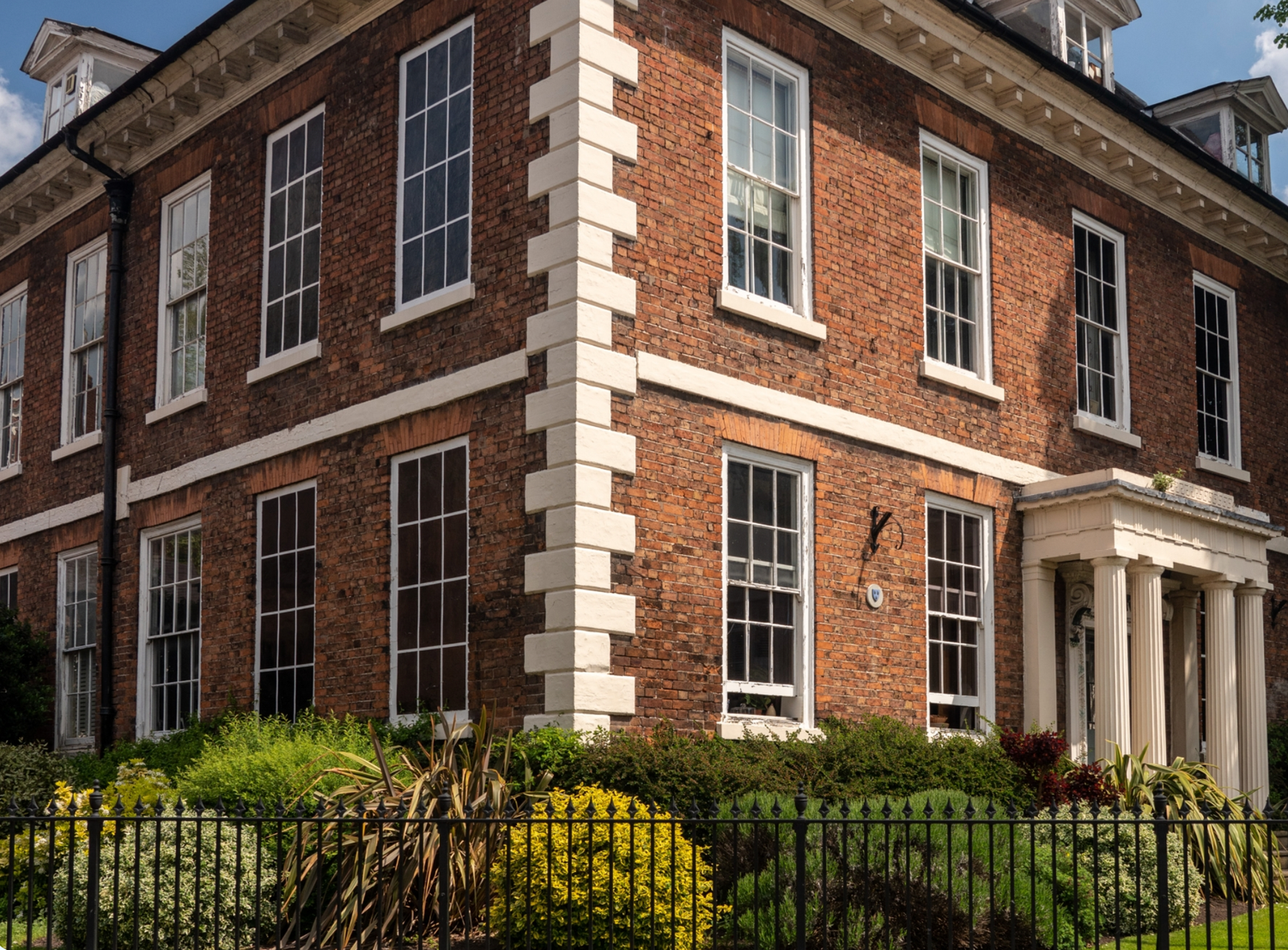Historic red brick Georgian manor house with white stone quoins, sash windows, and iron fence surrounded by yellow flowering shrubs.