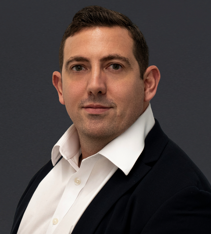 Professional headshot of a young man in a black blazer and white shirt against a gray background.