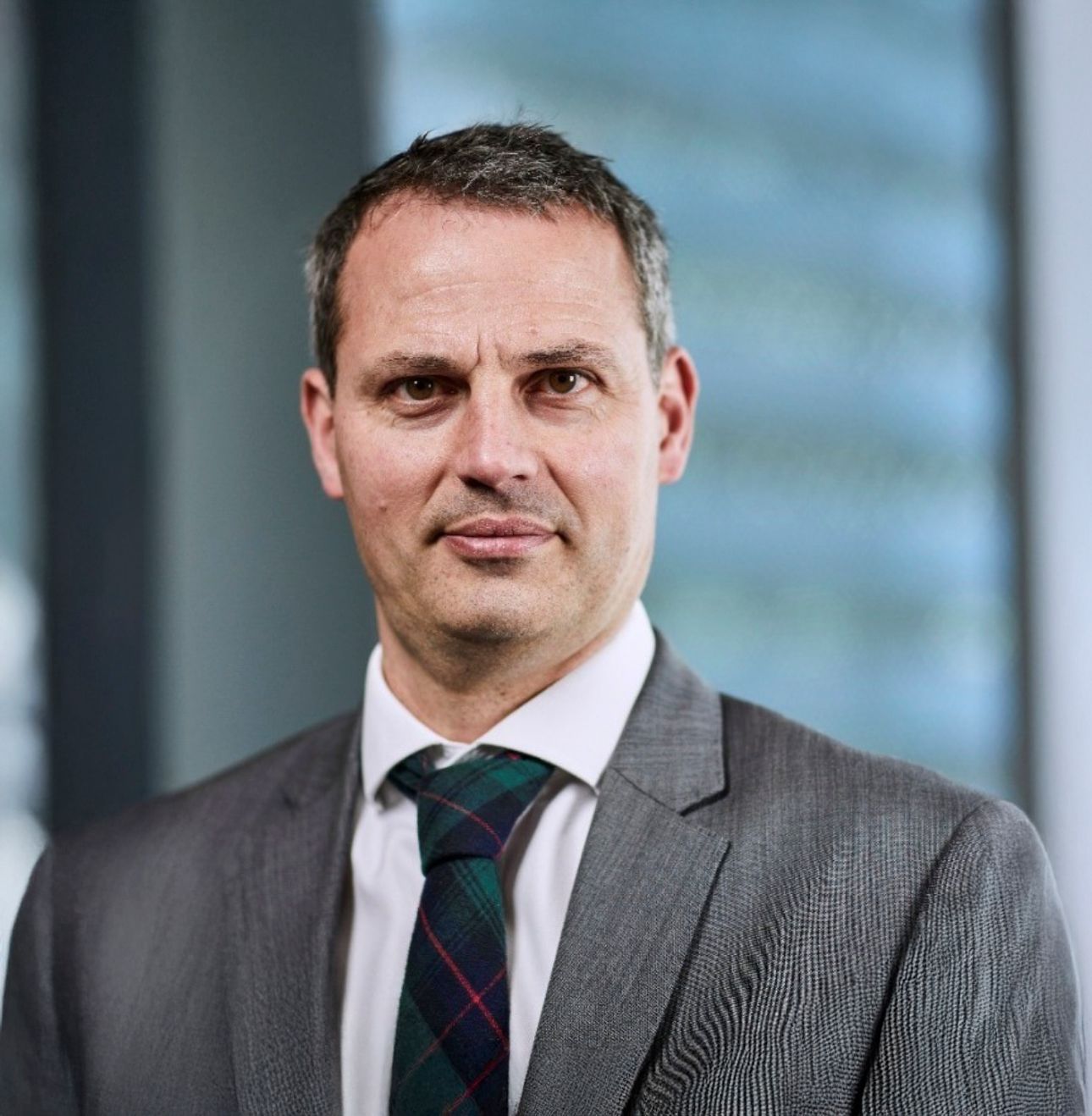 Professional businessman in gray suit and plaid tie posing for corporate headshot in modern office setting.