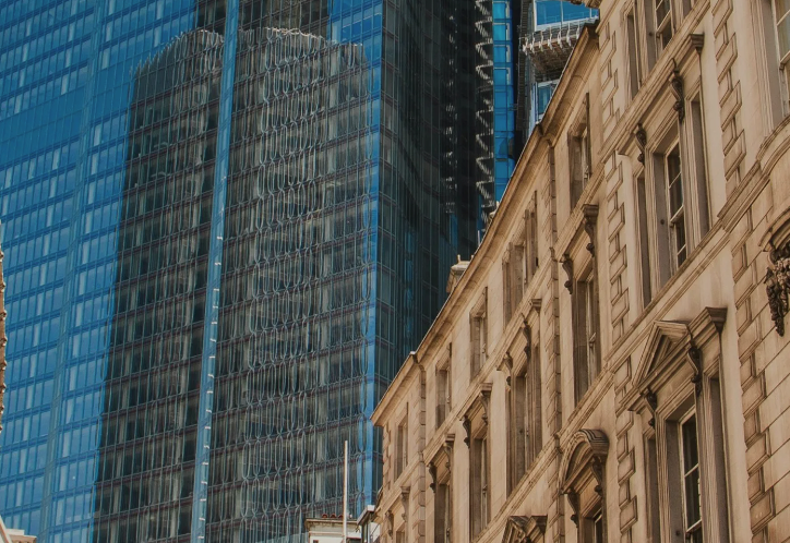 Modern glass skyscraper contrasts with ornate classical stone building facade in urban cityscape.
