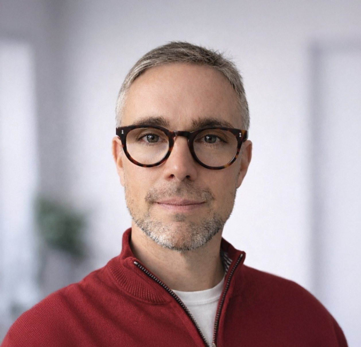 Middle-aged man with gray hair, black glasses, and a red zip-up sweater smiling in a bright, modern setting.