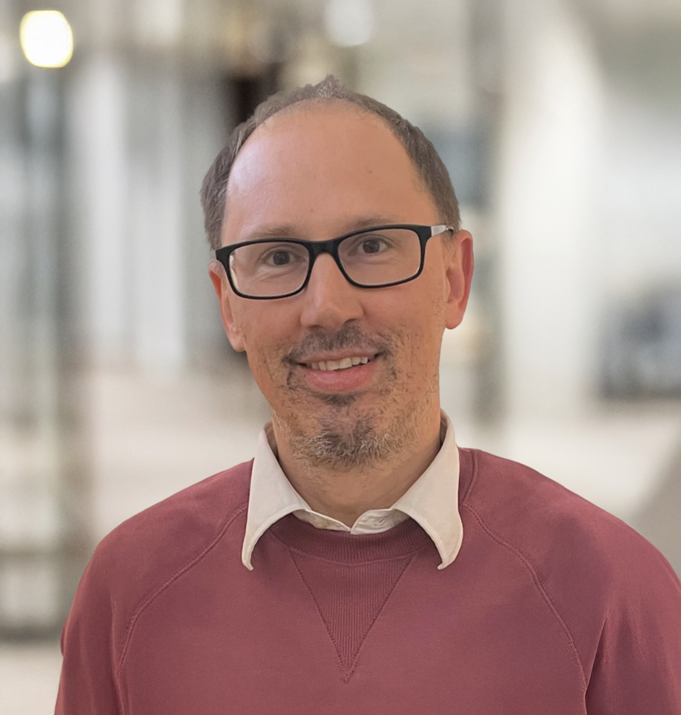 Smiling middle-aged man with glasses and goatee wearing a red sweater in a modern office setting.