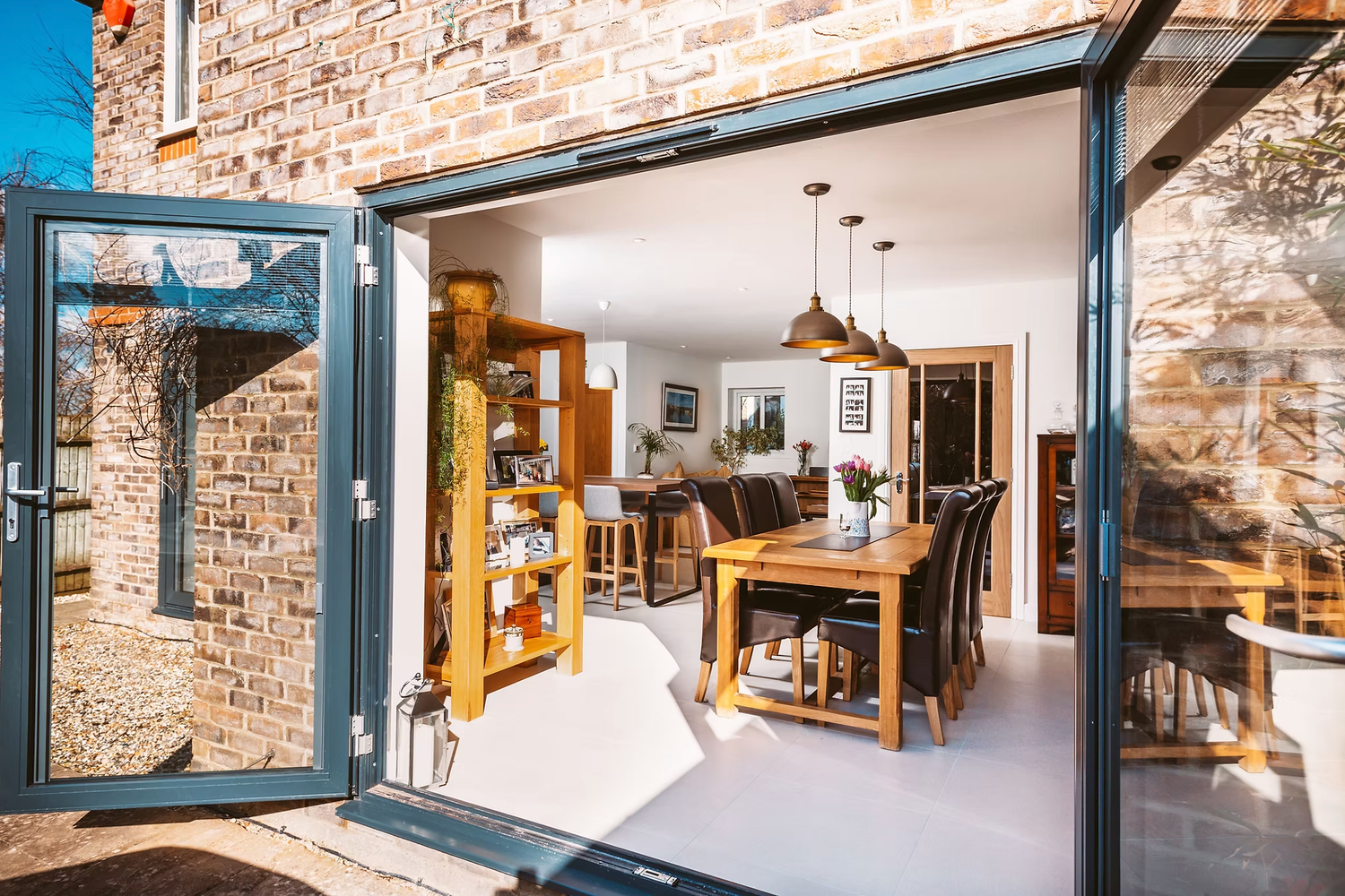 Bright dining room with wooden furniture viewed through modern bi-fold doors in stone cottage.