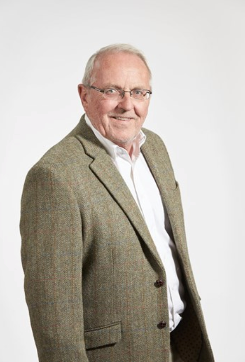 Smiling senior man with white hair and glasses wearing a green tweed blazer and white shirt against a light background.