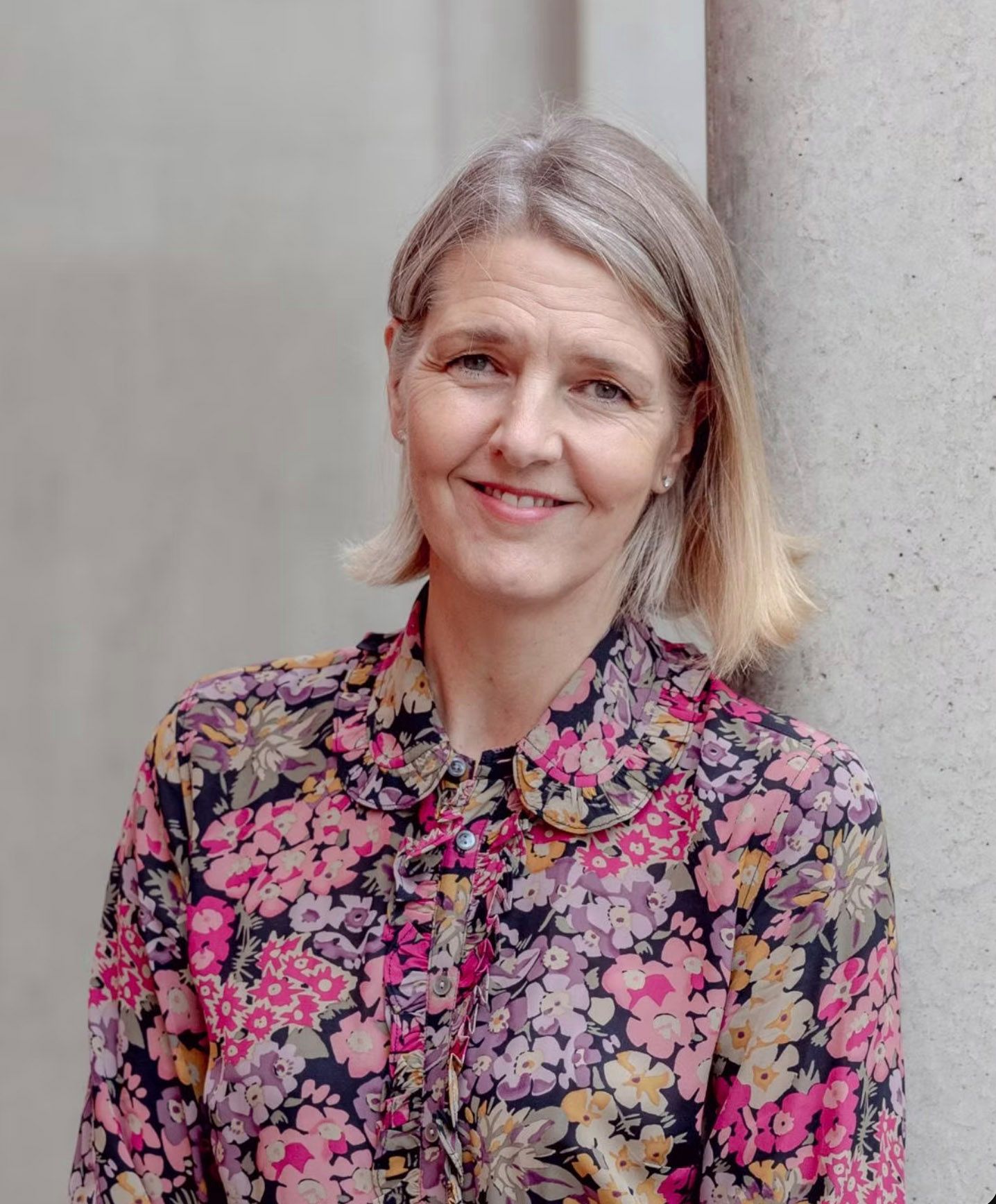 Smiling woman with blonde hair wearing a floral shirt, leaning against a column in a bright outdoor setting.