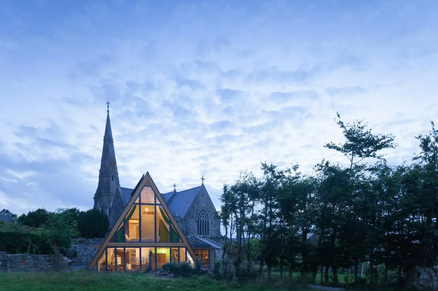 Modern A-frame wooden extension beside historic stone church with illuminated interior at dusk under dramatic blue sky.