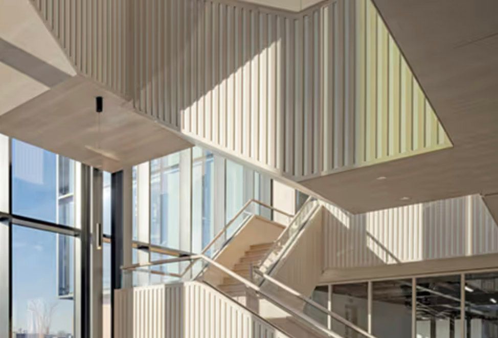 Modern building interior with geometric staircase, ribbed white walls, and floor-to-ceiling windows flooding the space with natural light.