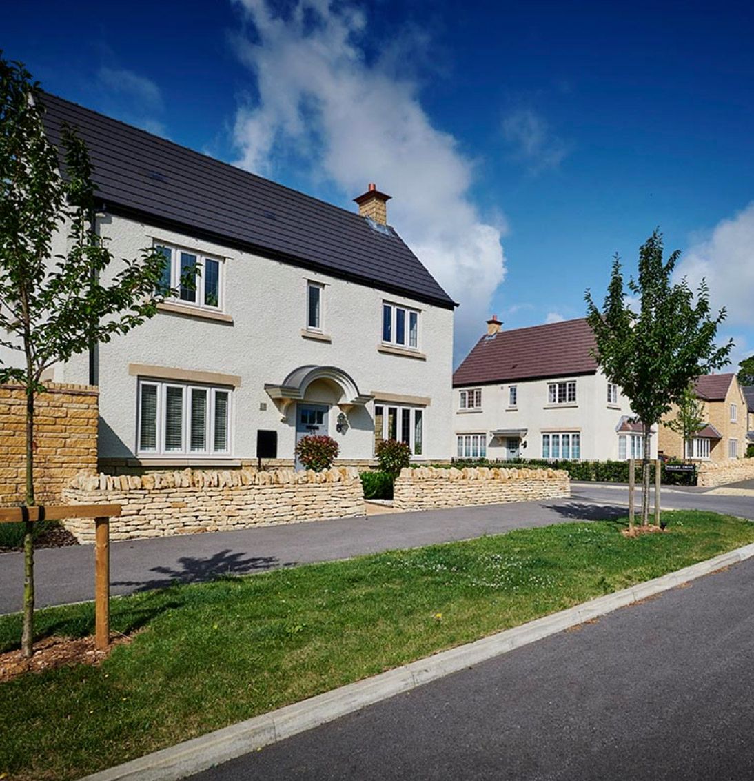 Modern Cotswold stone detached houses with slate roofs, manicured lawns, and young trees lining a quiet residential street.