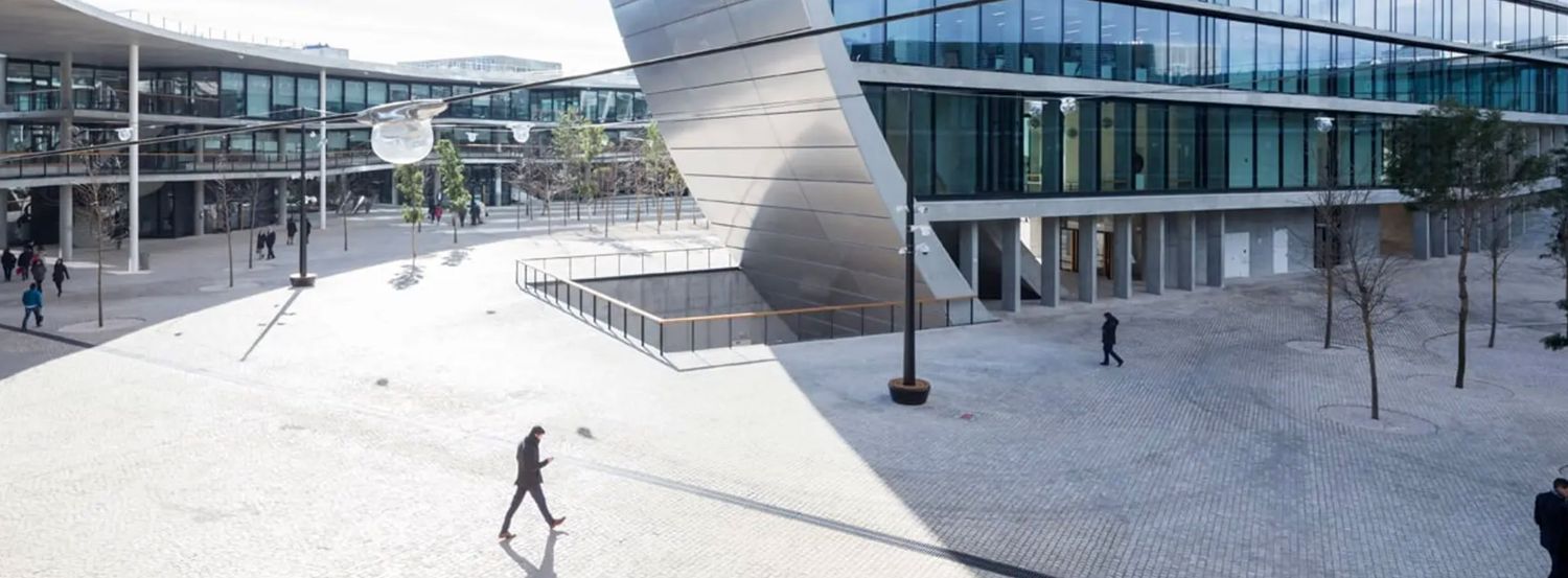 Modern architectural plaza with angular building facades, open concrete courtyard, and pedestrians walking under clear blue sky.