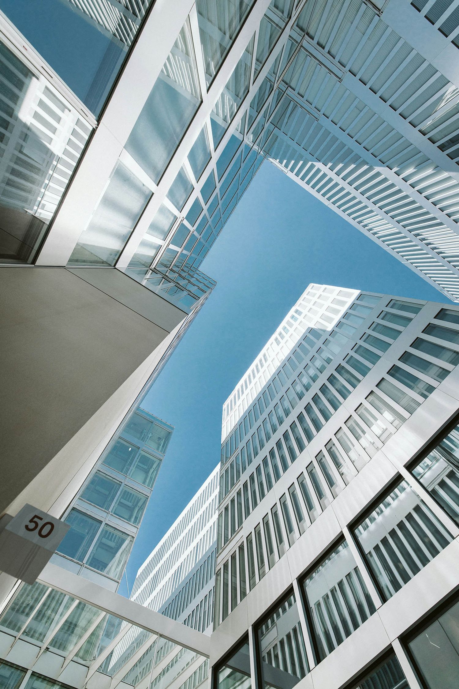 Looking up at modern glass skyscrapers converging toward a clear blue sky, with sleek white facades and reflective windows.