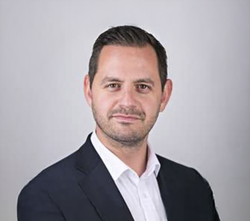 Professional businessman in dark blazer and white shirt poses for a corporate headshot against a gray background.