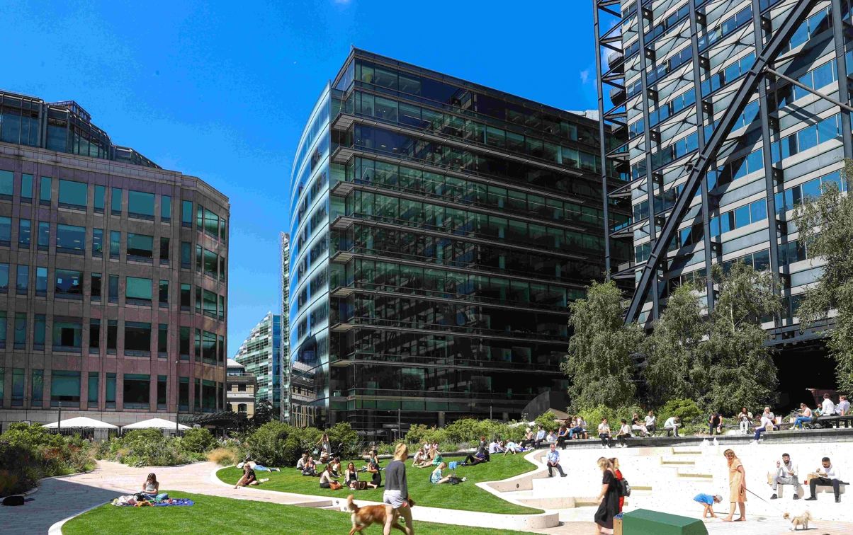Modern glass office buildings surround a vibrant urban park with people relaxing on green lawns under a clear blue sky.