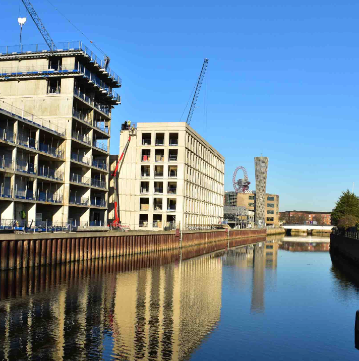Modern buildings under construction reflected in calm river water on a clear blue sky day.