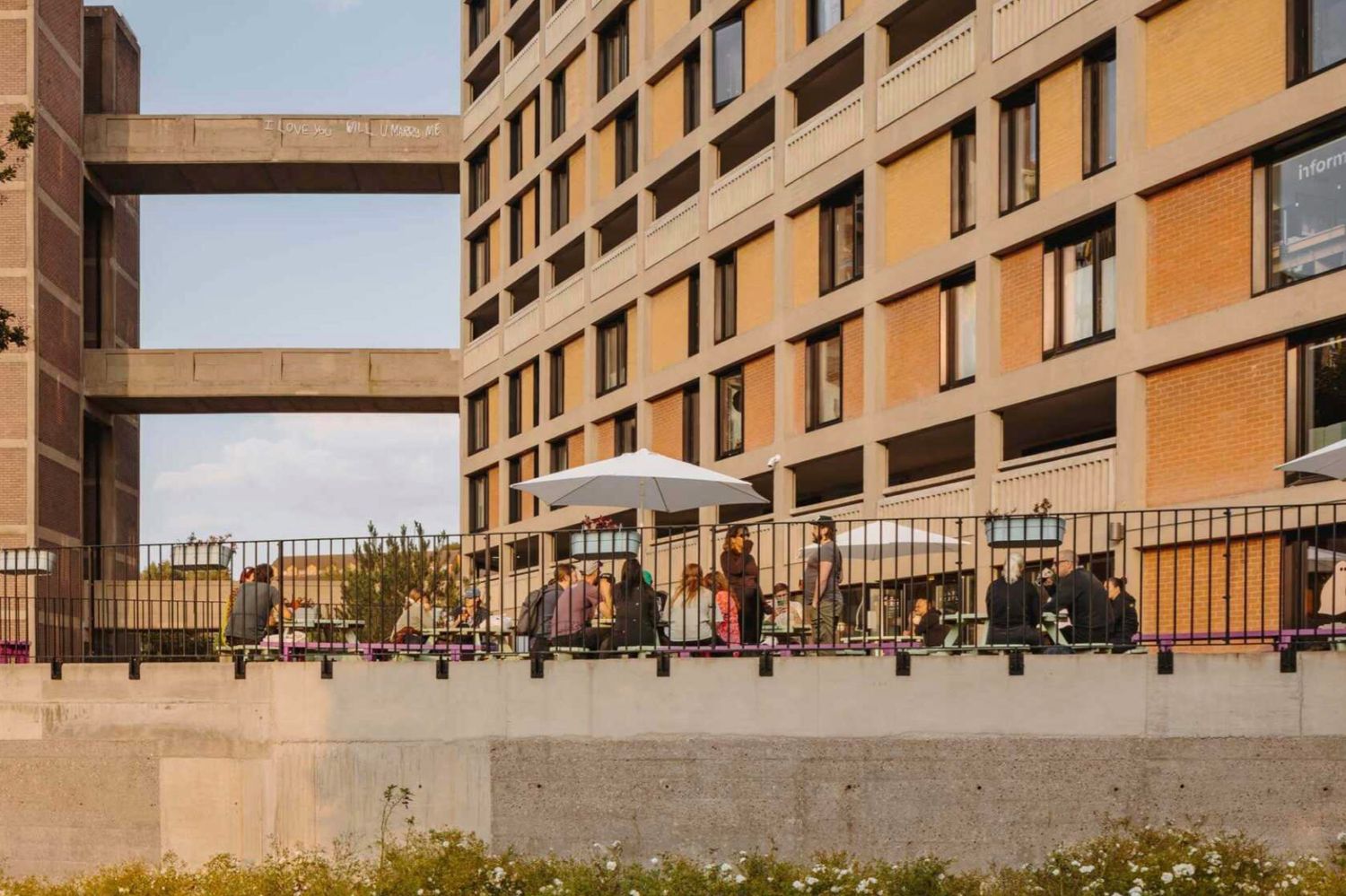 People gathered on a terrace of a modern brick building with large windows and an overhead walkway bridge.