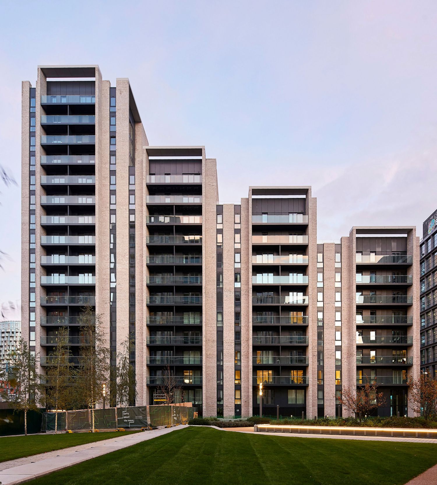 Modern high-rise residential apartment complex with balconies and dark brick facade surrounded by green lawn.