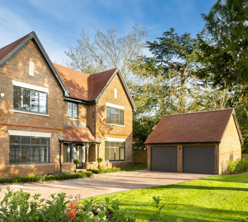 Large brick family home with red tile roof, double garage, manicured garden, and tree-lined backdrop on a sunny day.