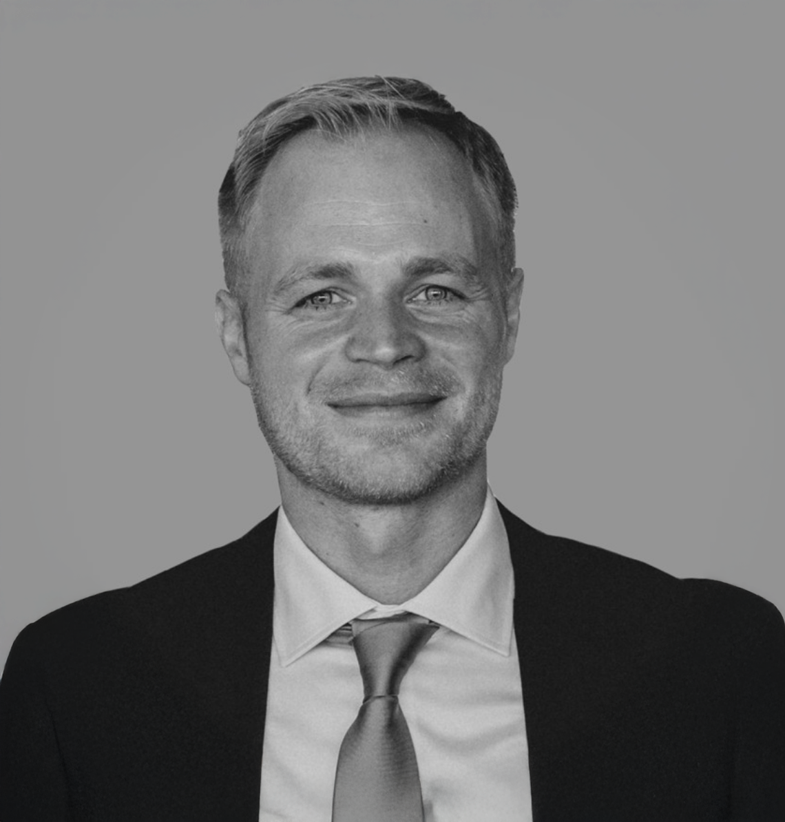 Professional black and white headshot of a smiling blond man in a dark suit and tie.