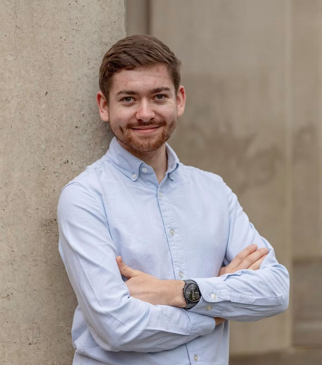 Young man with beard in light blue shirt, arms crossed, smiling confidently while leaning against a stone column.