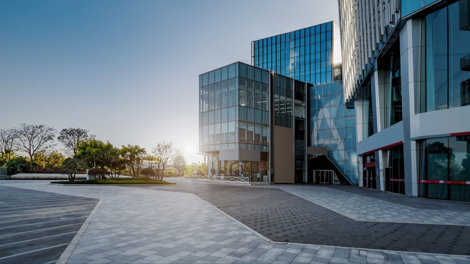 Modern glass office buildings with sunlight streaming through at golden hour on an open paved plaza.