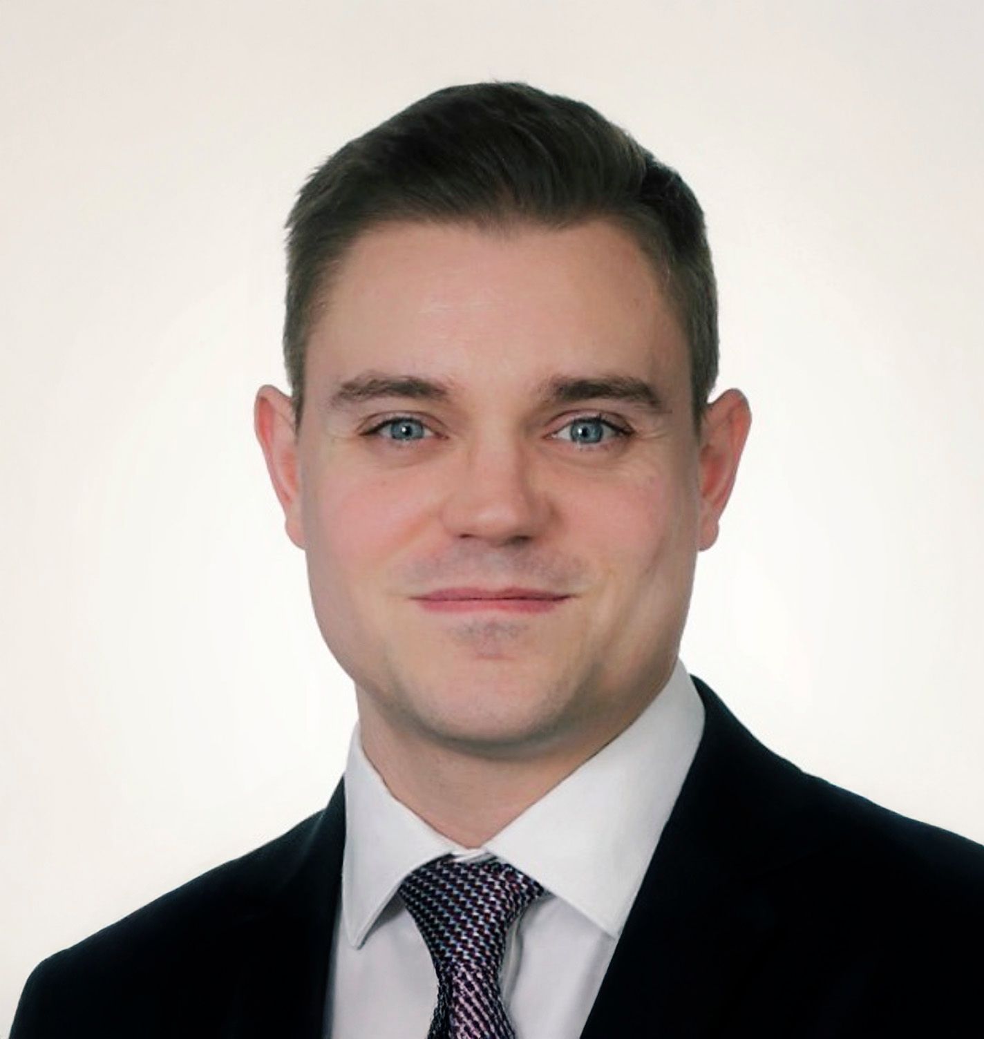 Professional headshot of a young man in a dark suit and patterned tie against a light background.