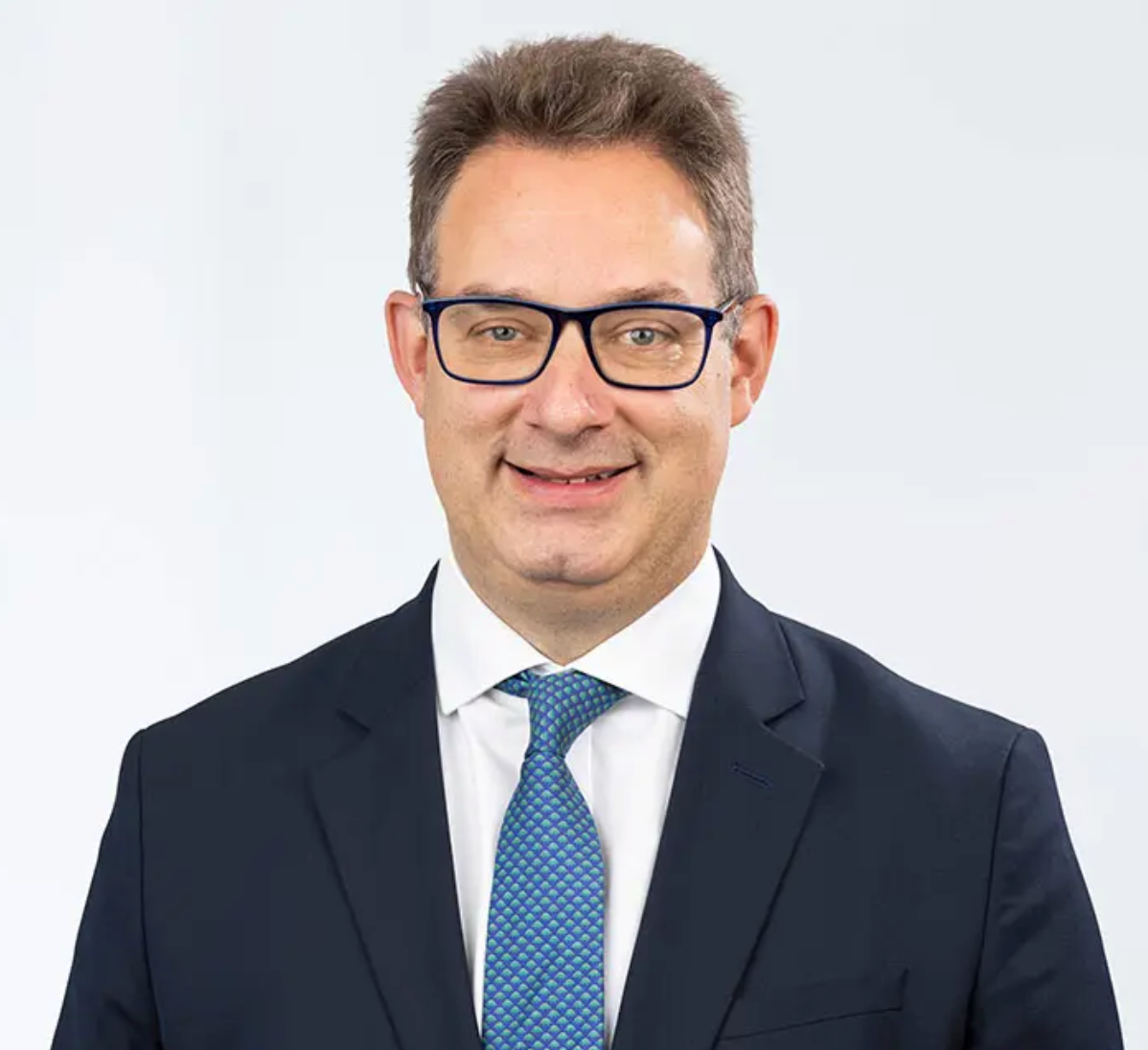 Professional businessman in dark suit and blue tie with glasses smiling against white background.