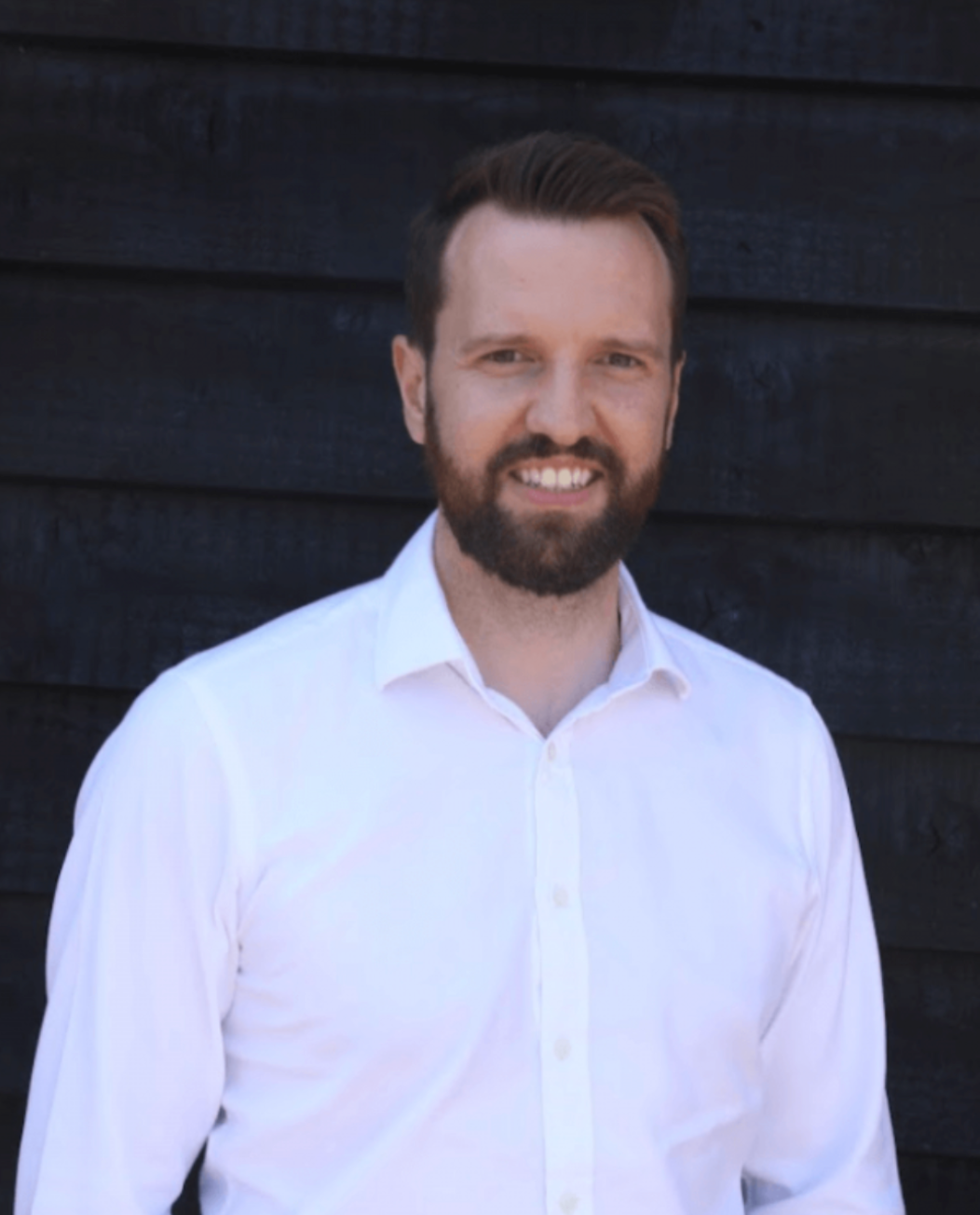 A smiling bearded man in a white dress shirt poses outdoors against a dark wooden wall background.