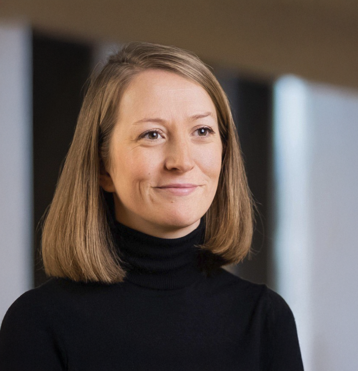 Professional headshot of a smiling woman with blonde bob hair wearing a black turtleneck sweater in a modern office setting.