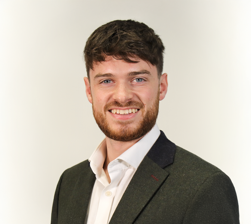 Young professional man with beard smiling, wearing dark green blazer and white shirt, studio portrait.