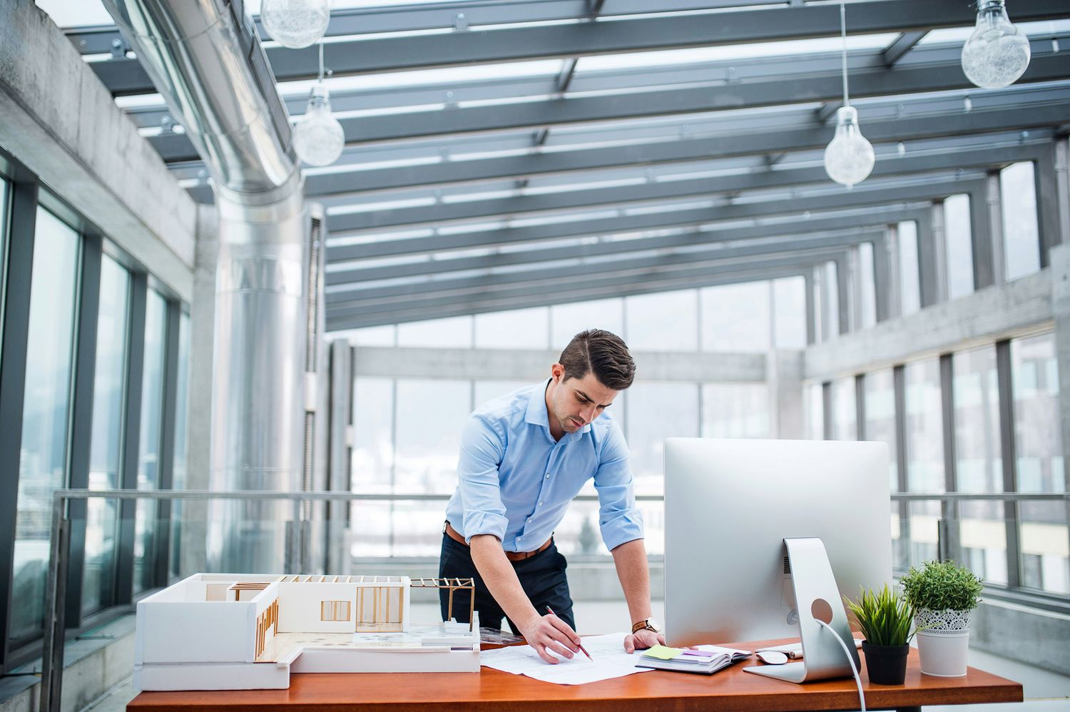 A professional man in a blue shirt stands at a modern desk reviewing documents next to a computer in a bright office.