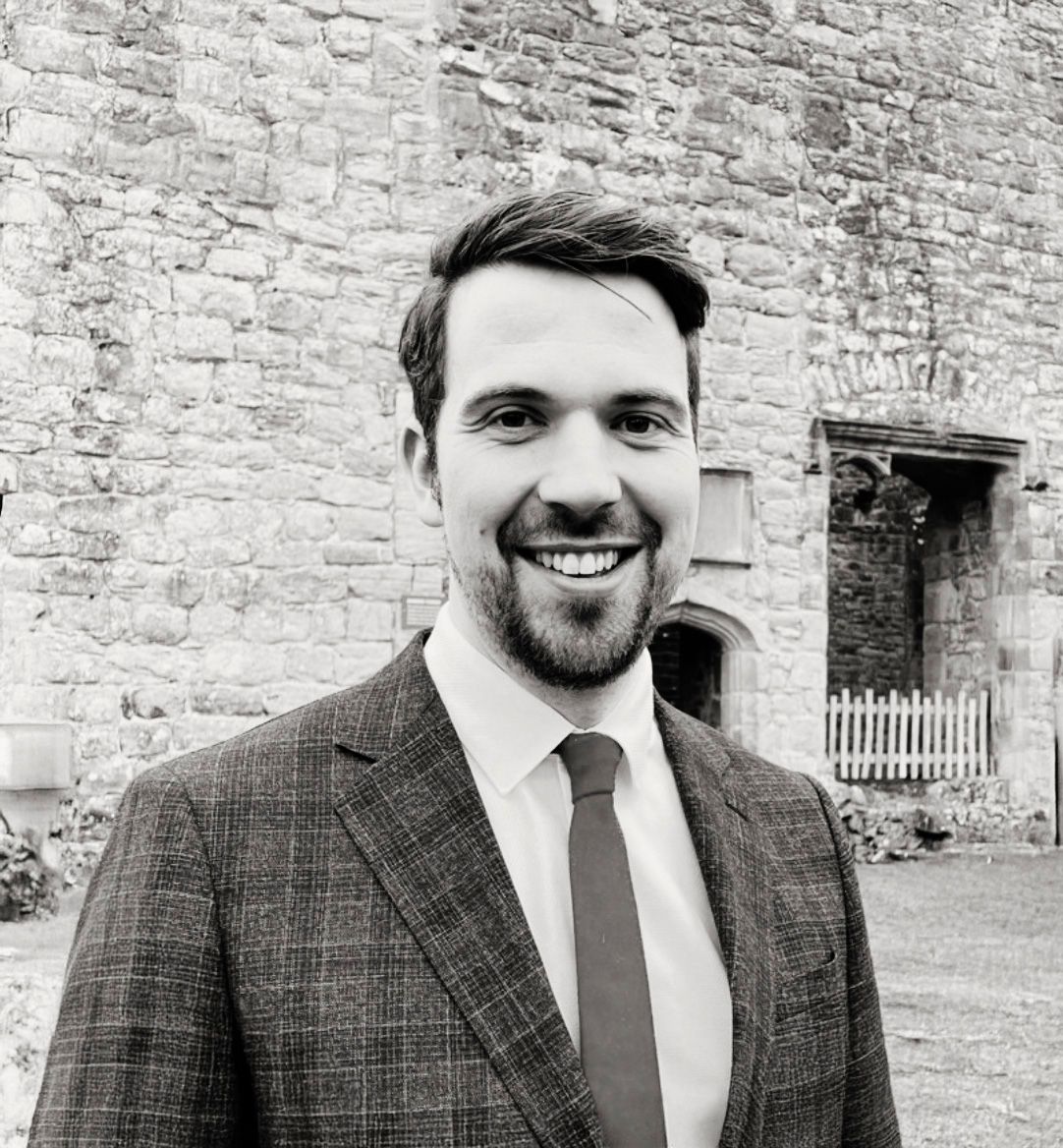 Smiling man with beard in plaid suit and tie standing before historic stone building outdoors.