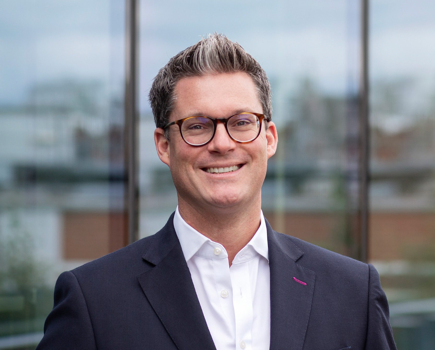 Professional businessman in dark blazer and glasses smiling confidently outdoors near modern building.