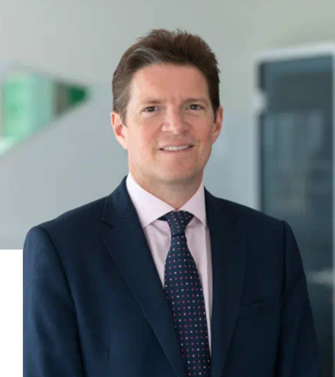 Professional businessman in navy suit and dotted tie smiling confidently in a modern office setting.