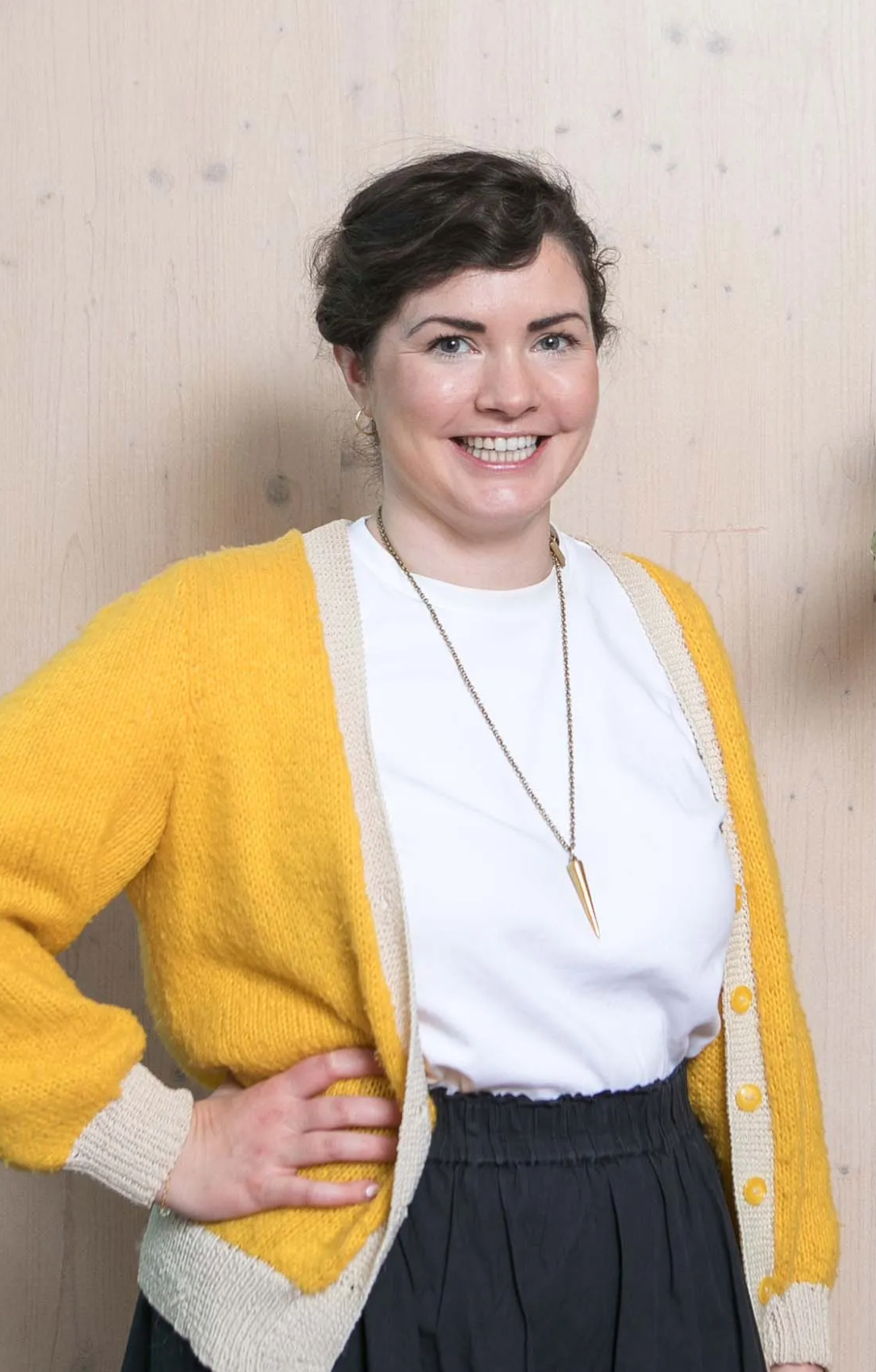 Smiling woman in yellow cardigan and white top with gold necklace posing against light wood background.