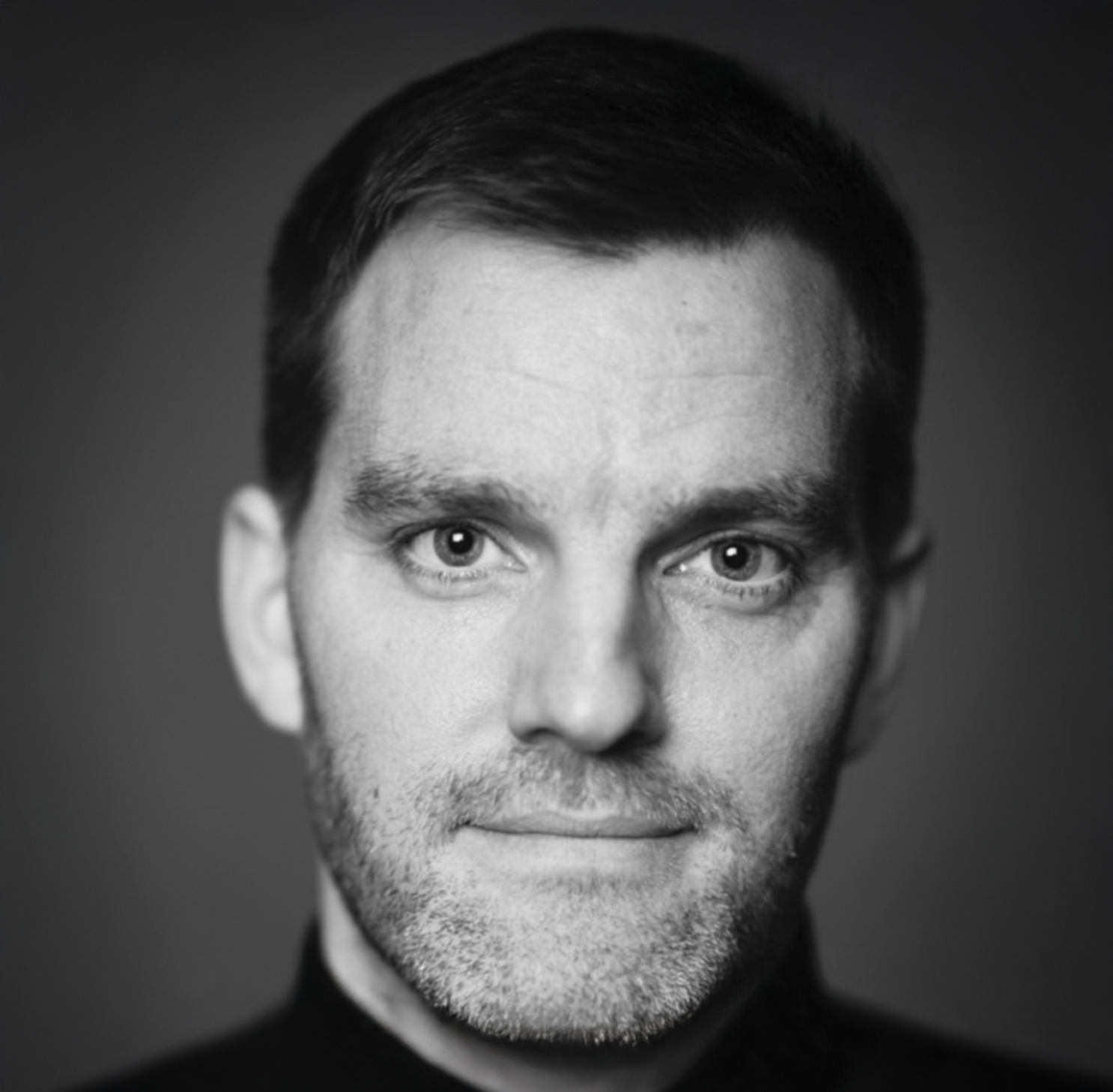 Black and white professional headshot of a man with short hair and light stubble wearing a dark turtleneck.
