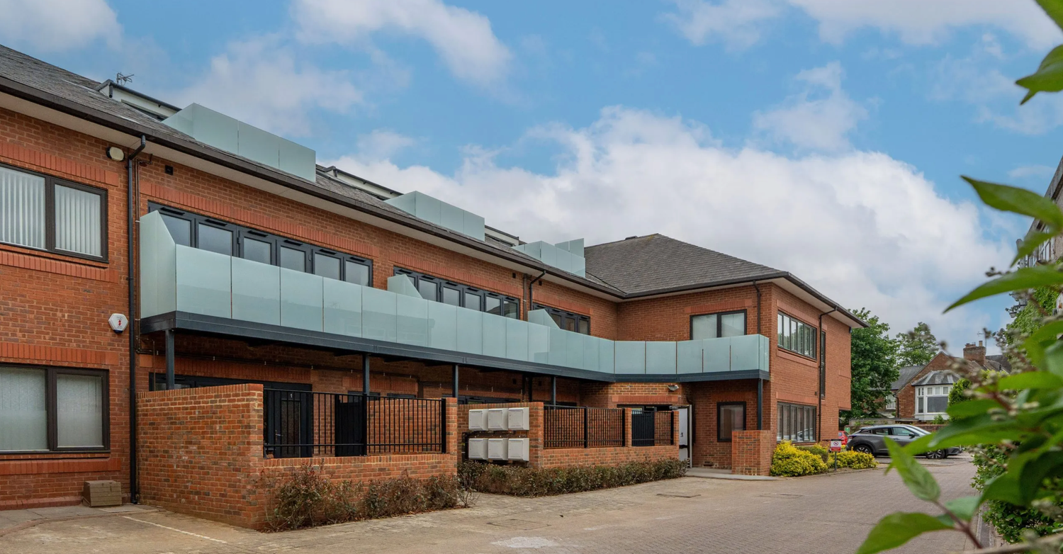 Modern two-story brick office building with glass canopy entrance, solar panels, and landscaped parking area under blue sky.