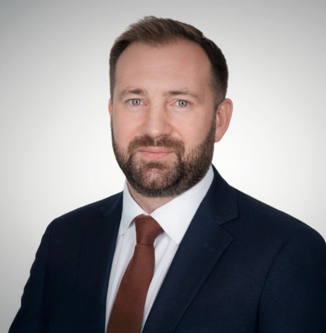 Professional headshot of a man in a navy suit and red tie with a neatly trimmed beard on a gray background.