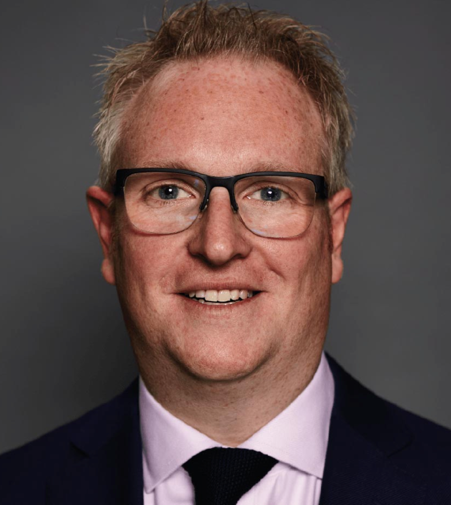 Professional headshot of a smiling middle-aged man with glasses, wearing a dark suit and tie against a gray background.