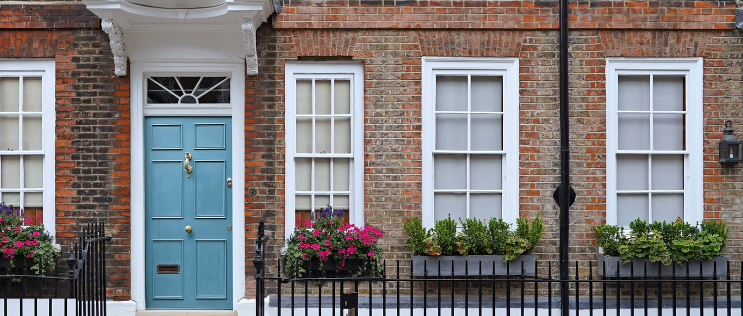 Charming brick townhouse facade with blue front door, white sash windows, and colorful window box flowers.