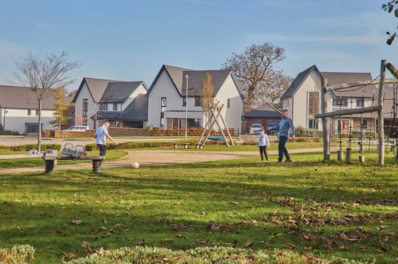 Families enjoy a green park area surrounded by modern suburban homes on a sunny autumn day.