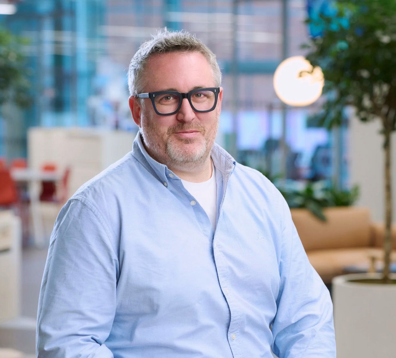 Middle-aged man with gray hair and black glasses wearing a light blue shirt in a modern office environment.