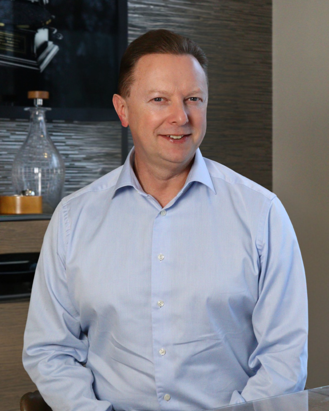 A smiling middle-aged man in a light blue dress shirt sits in a modern interior setting with decorative accents behind him.