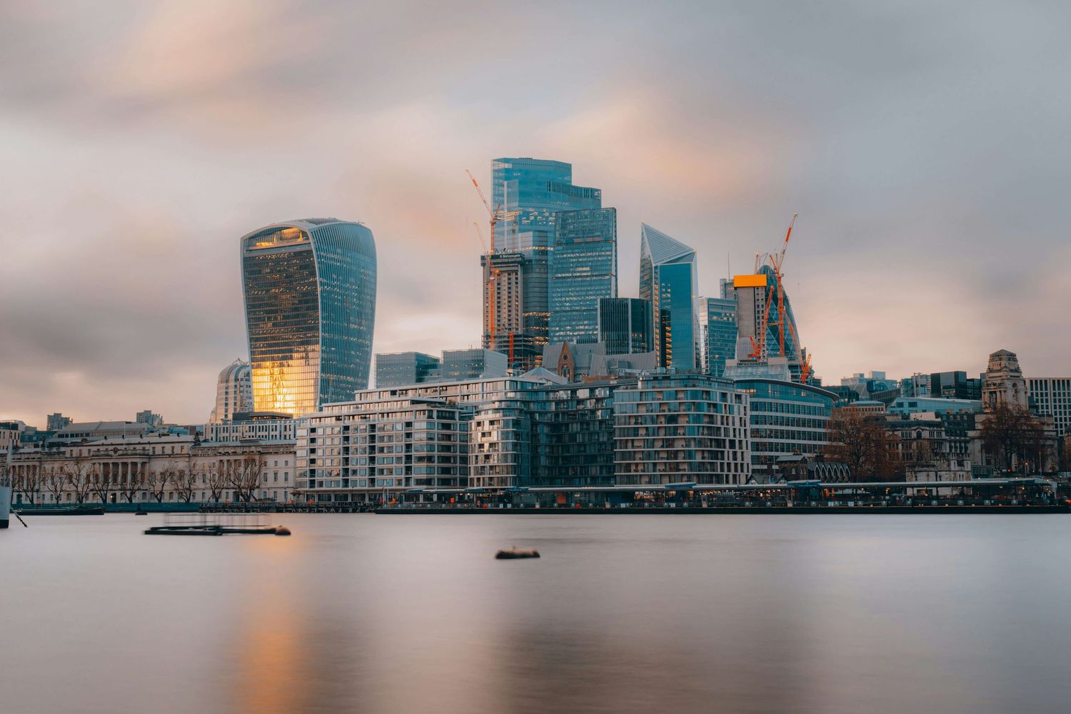 London City skyline with modern skyscrapers reflecting golden sunset light over the River Thames on a cloudy evening.