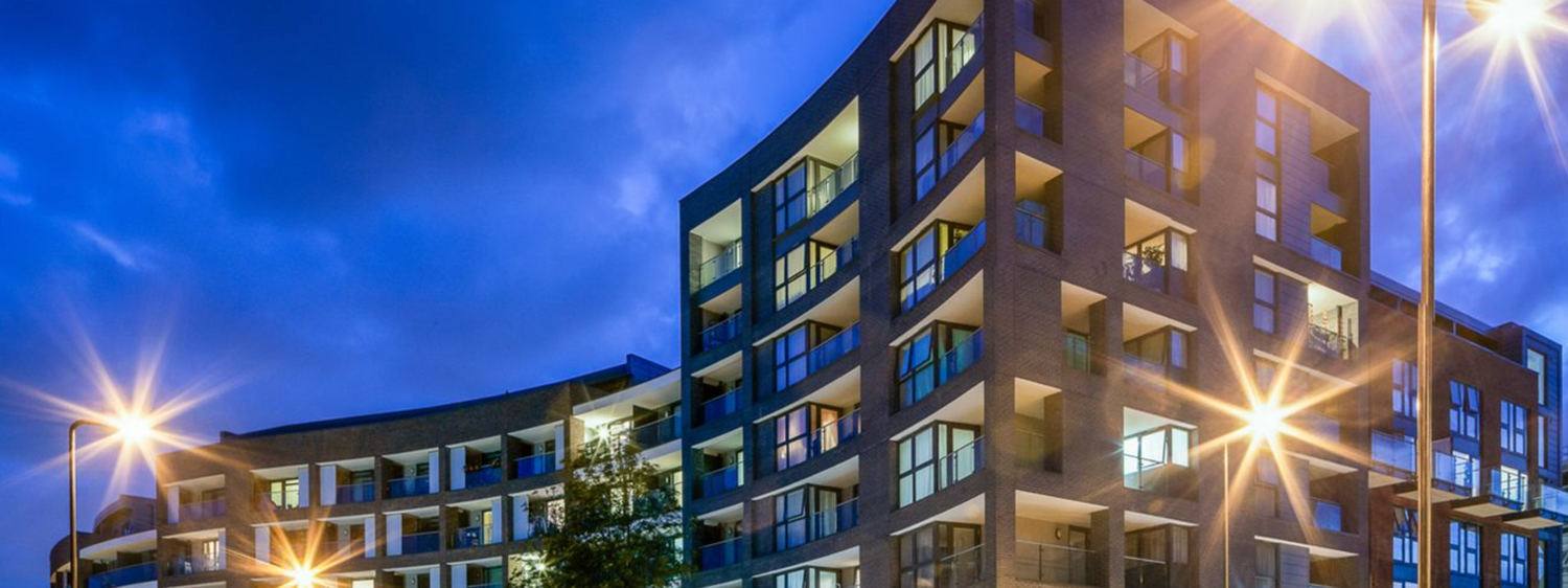 Modern apartment building illuminated at dusk with glowing windows against a dramatic blue evening sky.
