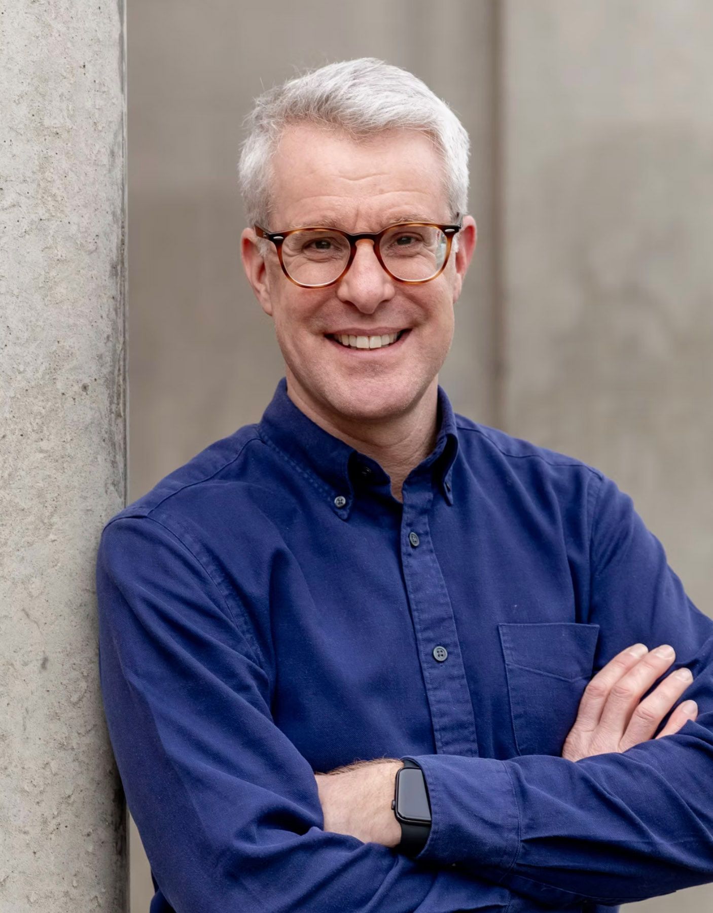 Smiling middle-aged man with gray hair and glasses, arms crossed, wearing a blue shirt, leaning against a concrete wall.