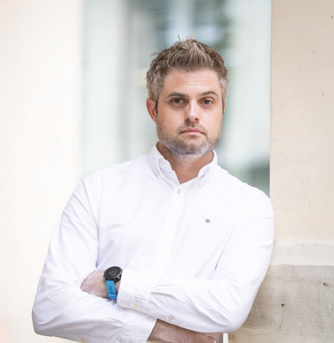 Professional man in white shirt with arms crossed, posing confidently against a modern architectural background.