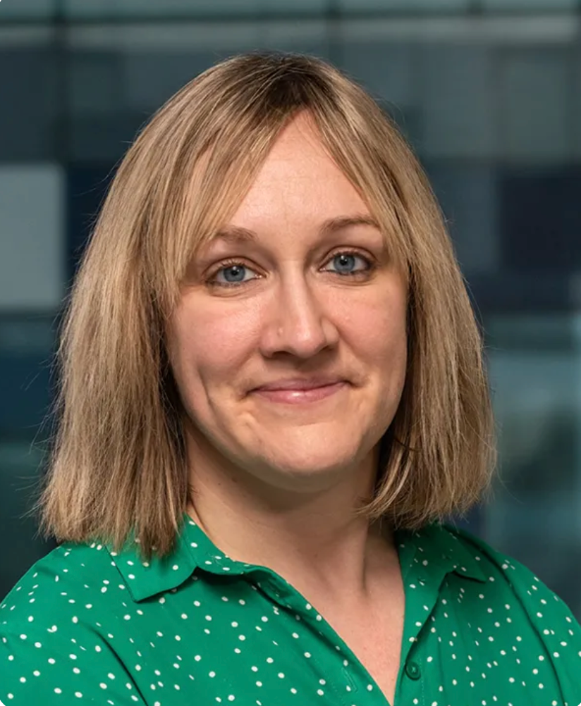 Professional headshot of a smiling woman with blonde hair wearing a green polka dot blouse in an office setting.