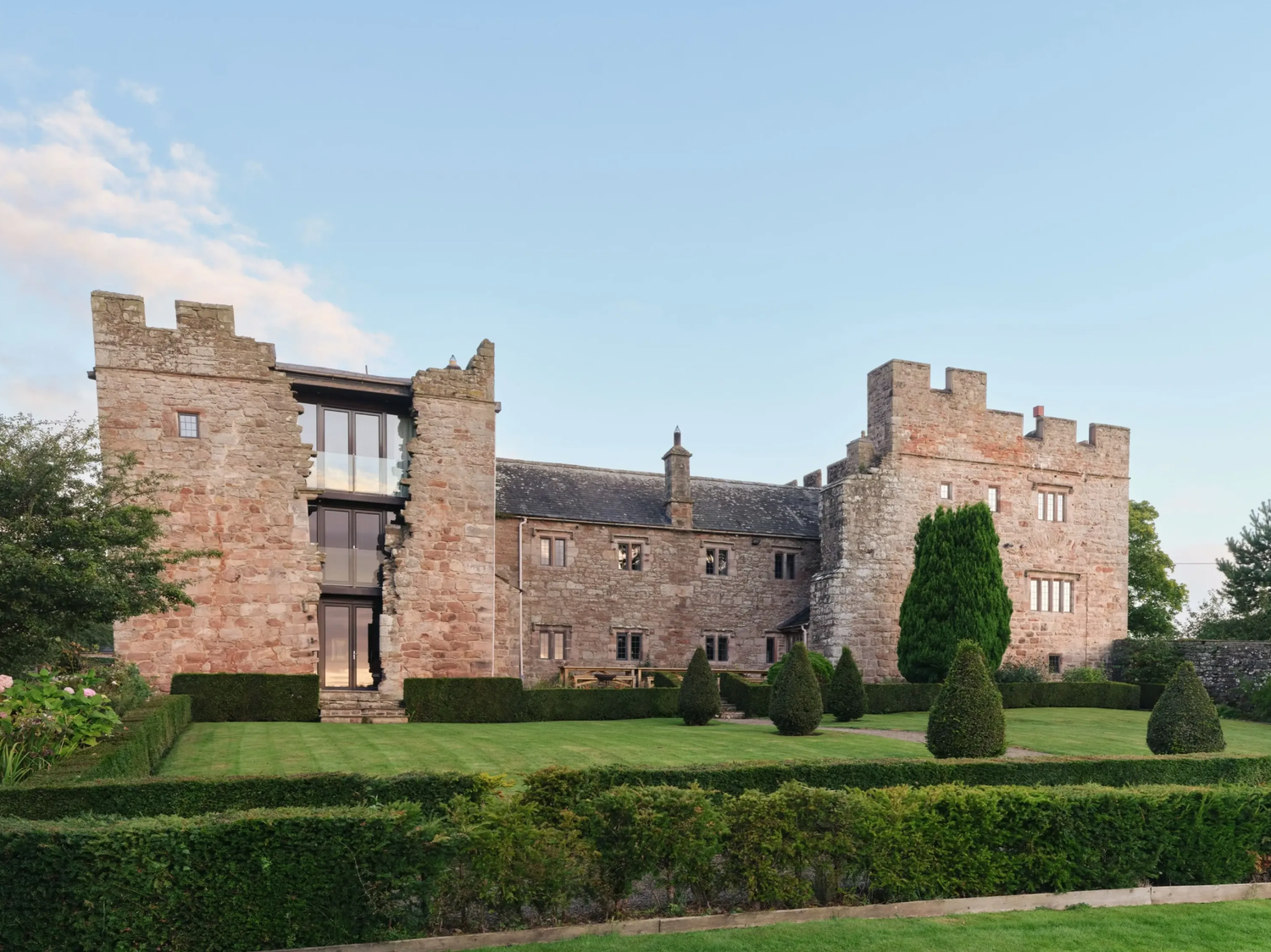 Historic medieval stone castle with manicured hedges, topiary trees, and lush green lawns under a clear blue sky.