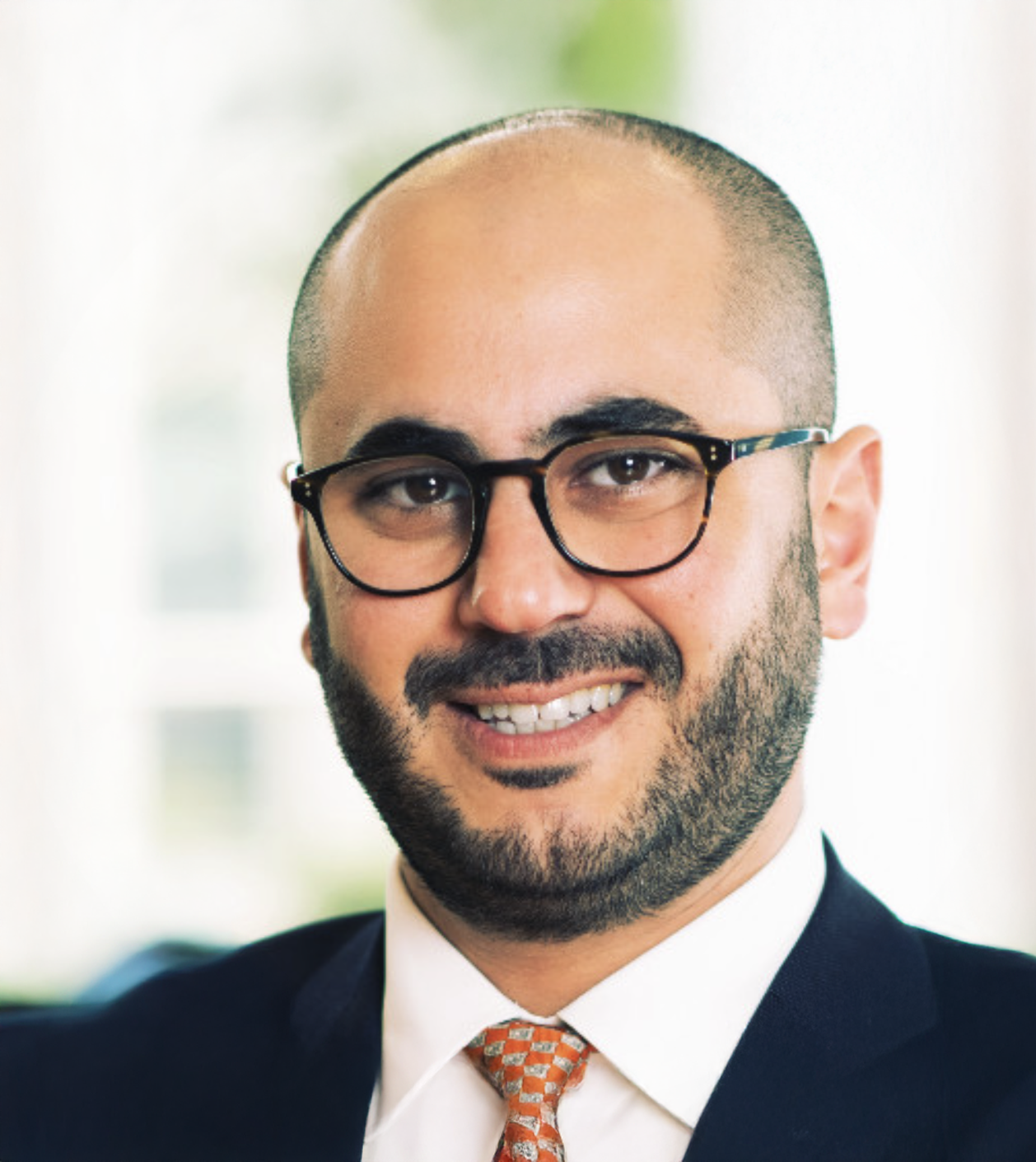 Professional headshot of a smiling man with glasses and a beard wearing a navy suit and orange tie.