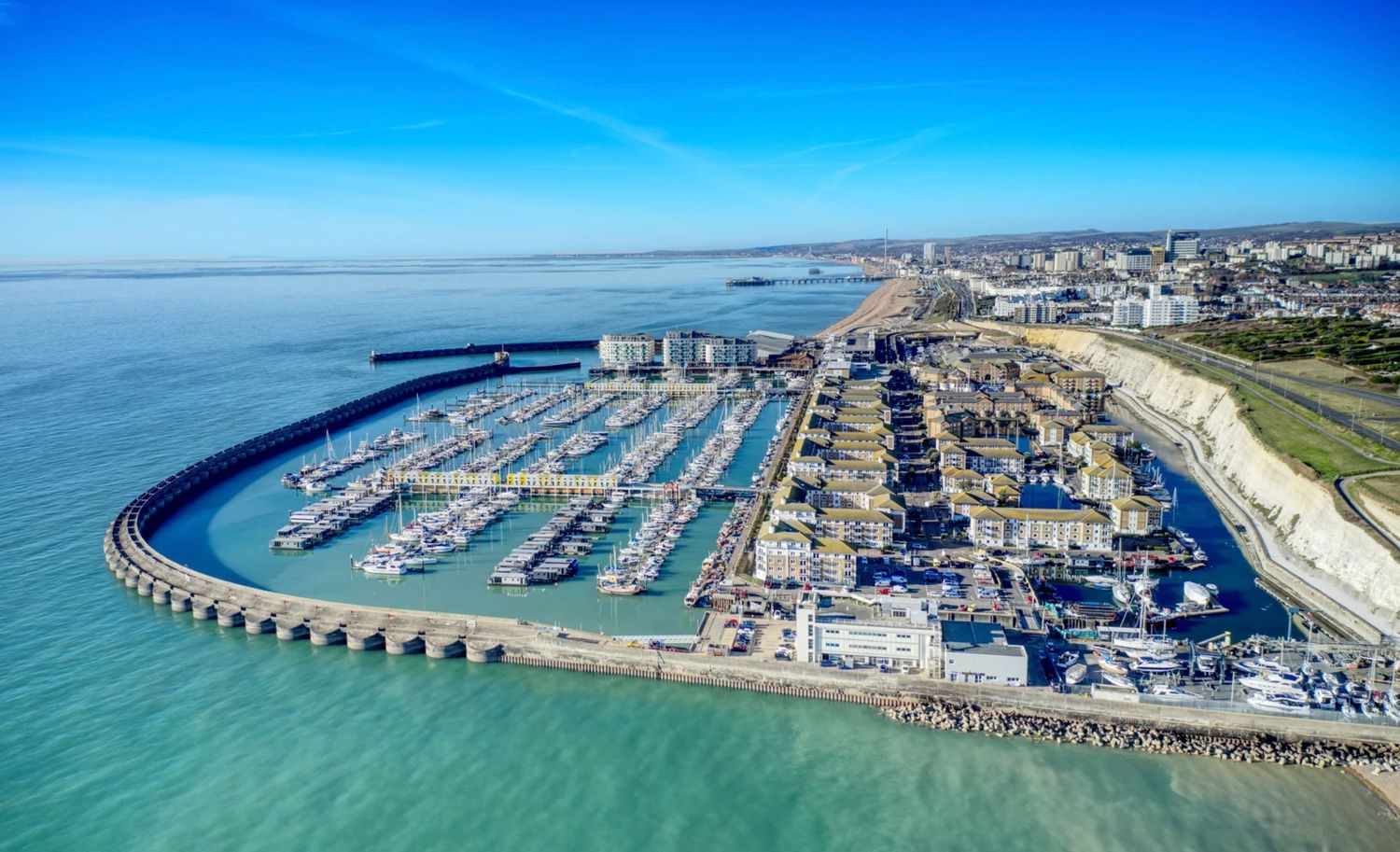 Aerial view of a bustling marina with boats, curved pier, and coastal city skyline under a clear blue sky.